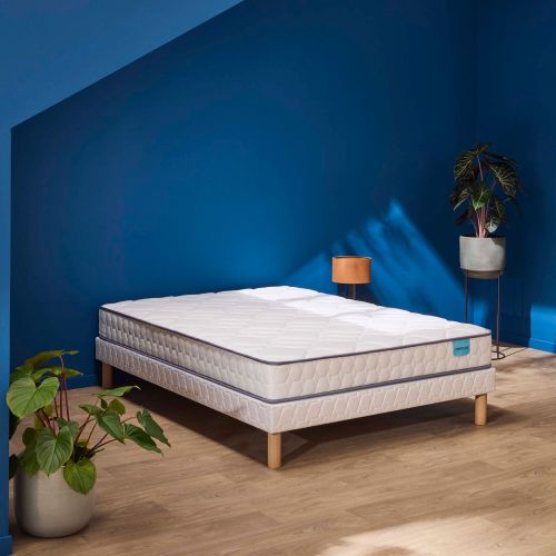 Matelas en mousse ferme Merinos Wise présenté sur un sommier dans une chambre au mur bleu.