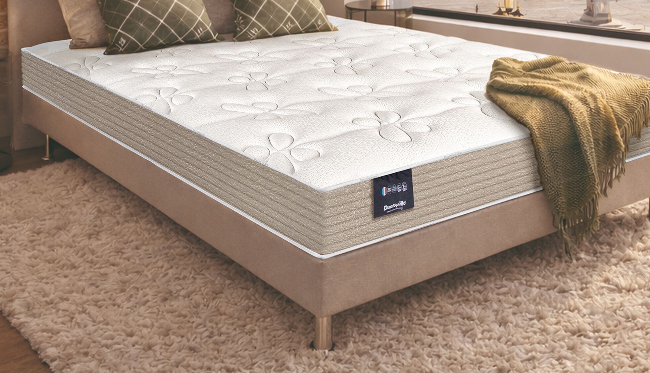 Matelas latex ferme Dunlopillo Dahlia 2 présenté en angle dans une chambre moderne avec briques.